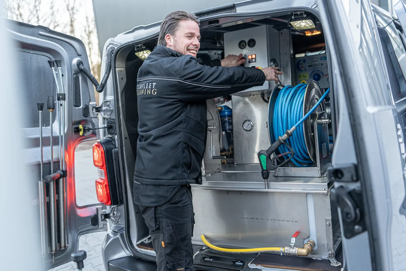 Lachende medewerker van Compleet Cleaning bedient de professionele hogedrukapparatuur in het bedrijfsbusje